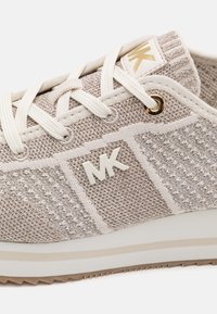 Chaussure de sport beige clair avec une texture tissée, des lacets blancs et un accent doré. Comprend un logo et une semelle rembourrée pour le confort.