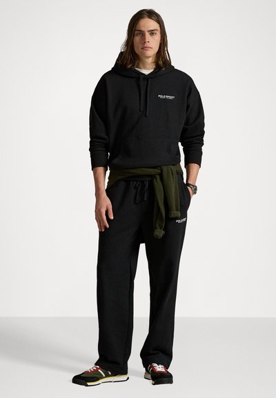 Hanorac negru cu glugă având un buzunar frontal și un logo, asociat cu pantaloni de trening asortați, un pulover verde legat la talie și adidași multicolori.