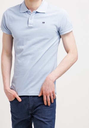 Homme portant un polo à manches courtes bleu clair et un jean bleu foncé, debout les mains près des poches devant un fond uni.