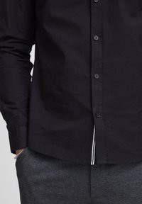 Mustassa nappikauluspaidassa on sileä kangas, klassinen kaulus ja hienovarainen valkoinen raita helman kohdalla. Yhdistetty harmaisiin struktuuripö trousers.