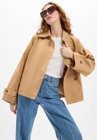 Lichtbruine jas met een kraag en grote knopen, gedragen over een wit shirt, gecombineerd met hoog uitgeknipte blauwe jeans.