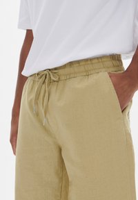 Beige drawstring-housut, joissa on elastinen vyötärö, sivutaskut ja kevyt, teksturoitu kangas. Yhdistetty valkoiseen paitaan.