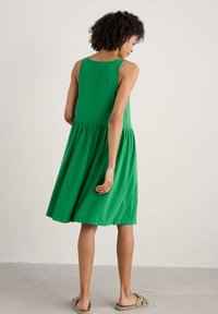 Groene mouwloze jurk met een getailleerde bovenkant en een geplooide rok. Gemaakt van zachte stof, valt hij net boven de knie. Platte sandalen maken de outfit compleet.