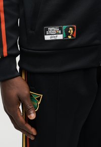 Tumša āda roka atpūšas pie melnām biksēm ar Jamaikas futbola emblēmu; melnā džemperī ir uzšuvums ar uzrakstu “Futbols ir brīvība” un Boba Mārlija attēlu.