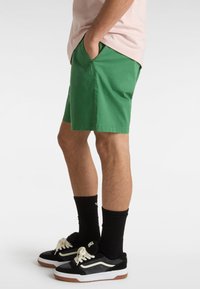 Grønne bomulds shorts med sidelommer, parret med sorte sneakers med hvide accenter og sorte sokker. Shortsene har en glat tekstur.