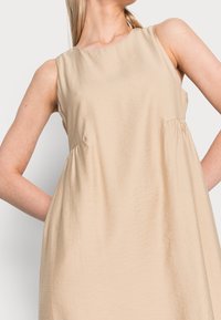 Béžové bezramínkové šaty s kulatým výstřihem, s plisovanými detaily na stranách a lehkým texturovaným materiálem.