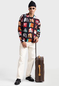 Kolorowa koszula z graficznym nadrukiem na czarnym tle, białe spodnie, czarne buty oraz brązowa walizka z monogramem i jasnobrązowymi akcentami.