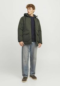 Dunkelgrüne gepolsterte Jacke mit Kapuze, Fronttaschen und Reißverschlussdetails, kombiniert mit einem navyblauen Pullover und hellblauen Jeans.