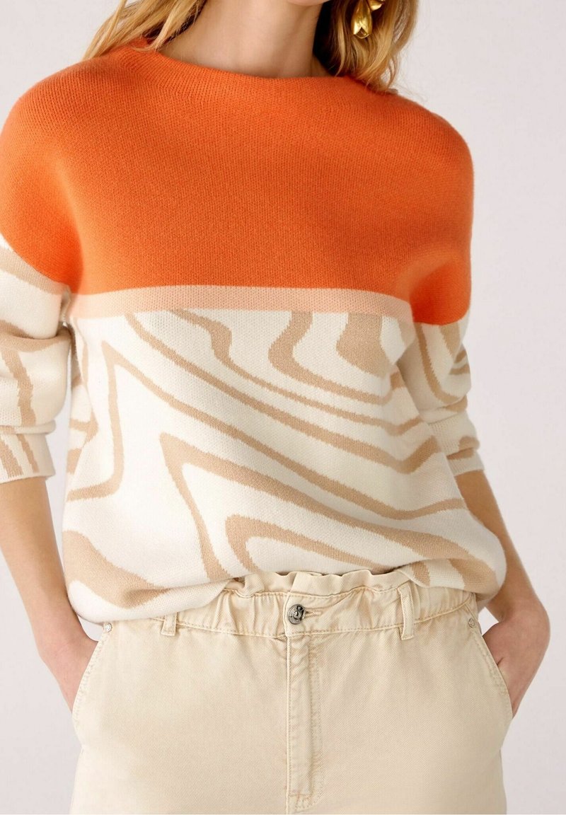 Maglione lavorato a maglia con parte superiore arancione, corpo crema con motivi astratti beige e polsini a coste. Pantaloni beige a vita alta visibili sotto.