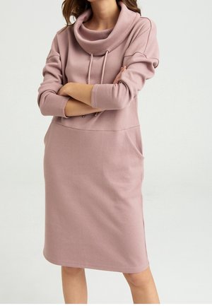 Mujer con un vestido rosa empolvado hasta la rodilla con cuello vuelto, mangas tres cuartos y bolsillos laterales, de pie con los brazos cruzados.