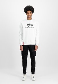 Bijela dukserica s crnim logom, uparen s crnim cargo hlačama s bočnim džepovima. Model nosi bijele tenisice s šarenim akcentima.