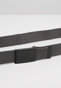 Grå vævet bælte med en sort metalspænde. Har et logo på spænden og en tekstureret finish. Justerbar længde for pasform.