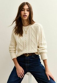 Sweter w kolorze kremowym, pleciony w warkocz, z okrągłym dekoltem, prążkowanymi mankietami i teksturowanymi wzorami, w zestawieniu z ciemnoniebieskimi dżinsami i brązowym paskiem.