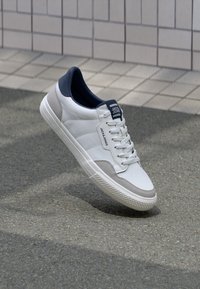 White sneaker with fabric and suede accents. Features a rubber sole, contrasting navy collar, and a logo tag on the side.