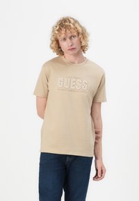 Beige bomull T-skjorte med korte ermer som har "GUESS"-logoen i hevet hvit tekst, rund hals, tettsittende design og glatt tekstur.