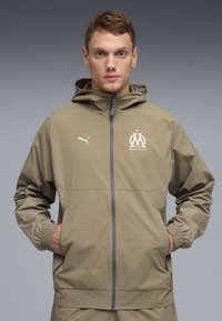 Ανοιχτό μπεζ μπουφάν με κουκούλα και ελαστικές μανσέτες, με κλείσιμο με φερμουάρ και λογότυπο στο στήθος. Κατασκευασμένο από λείο, αδιαβροχοποιημένο ύφασμα.