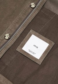 Tessuto marrone della giacca con cerniera argentata, due bottoni e un'etichetta bianca cucita all'interno che recita "MNG, EUR-USA M MEX M, Made in India."