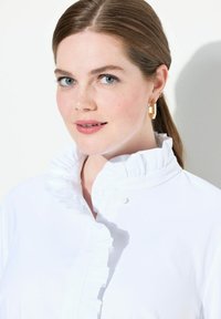 Camicia bianca con colletto alto e volant e chiusura a bottoni. Il modello indossa orecchini geometrici dorati, caratterizzati da una texture liscia e colori tenui.