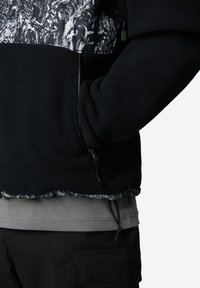 Czarna polarowa kurtka z wzorzystą szarością górną częścią, z pół-zamkiem i regulowanym dołem. Nosi się na szarej koszulce.