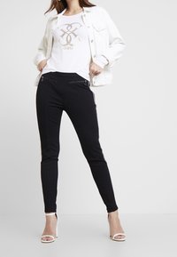 Giacca in denim bianco, top bianco con logo dorato, pantaloni neri aderenti con tasche con zip e tacchi a stiletto bianchi.