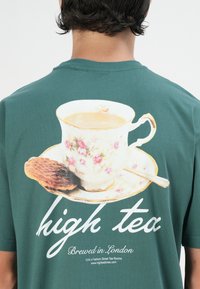 Zaļa T-krekls ar vintage tējas krūzi ar ziedu rakstu, šķīvīti un cepumi, kā arī ar tekstu "augstās tējas" un "Pagatavots Londonā."