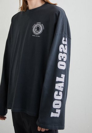 Person trägt ein schwarzes Langarmshirt mit weißem "LOCAL 032C" Schriftzug am Ärmel und einem kleinen runden Logo mit Text auf der Brust.