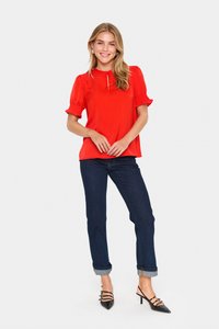 Top corto rosso con maniche a sbuffo e scollatura a chiave, abbinato a jeans blu scuro con risvolti e décolleté nere a punta.