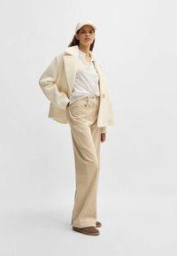 Cappotto in tessuto cremoso, camicia a pois bianchi, pantaloni beige e sneakers marroni. Il cappello abbinato è dello stesso colore del cappotto. Design semplice e casual.