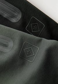 Dve panely textilného materiálu, jeden tmavosivý a jeden čierny, s embosovanými logami a predĺženými otvormi pre jednoduché prenášanie.