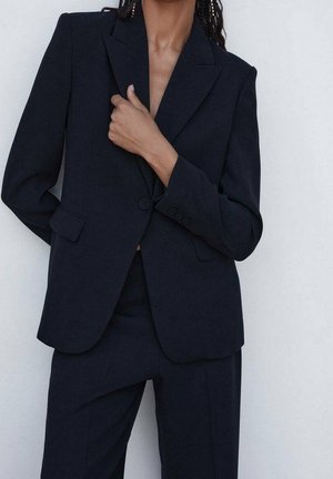 Personne portant un costume noir sur mesure avec une veste à un bouton et un pantalon assorti, la main reposant près du col de la veste.