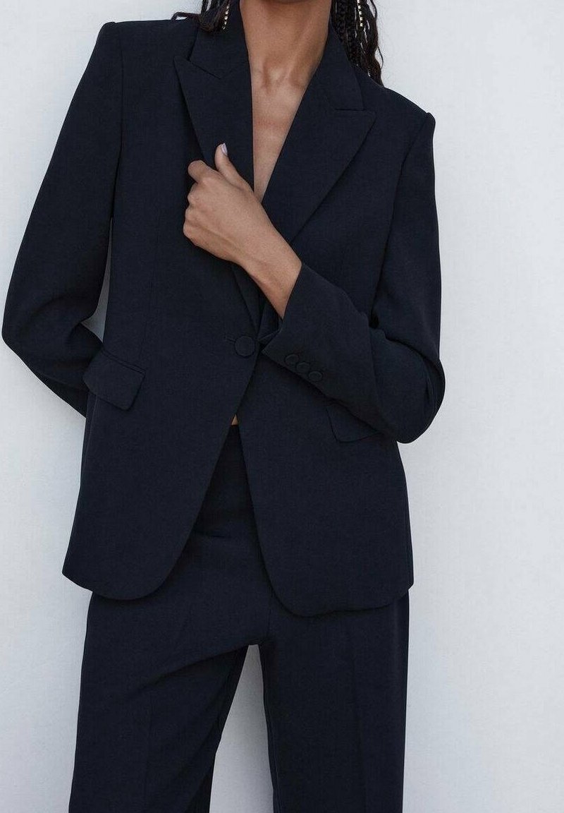 Personne portant un costume noir sur mesure avec une veste à un bouton et un pantalon assorti, la main reposant près du col de la veste.