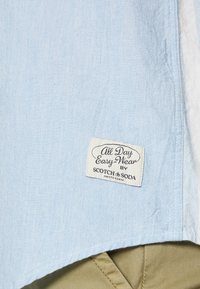 Vaaleansininen lyhythihaoinen paita, joka on valmistettu pehmeästä kankaasta. Paita sisältää "All Day Easy-Wear" -tarran tuotemerkiltä Scotch & Soda ja siinä on ommellut helma yksityiskohdat.