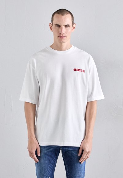 T-shirt bianco a maniche corte in cotone, con una stampa rossa "OBSESSED2" sull'area sinistra del petto. Indossato con jeans blu sfilacciati.