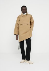 Pulover impermeabilizat bej cu buzunar frontal, șnururi reglabile la tiv și mâneci largi, asortat cu pantaloni negri și pantofi crem.