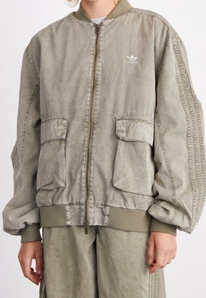 Person trägt locker sitzende, ausgewaschene olivgrüne Adidas Bomberjacke mit Fronttaschen und passende Hose, Reißverschluss geschlossen.