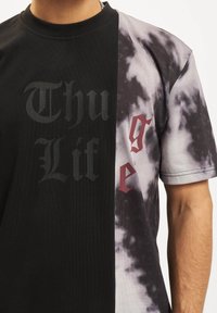 Puse melna, puse pelēka tīrīta īsā piedurkne T-kreklu ar uzrakstu "Thug Life" melnā un sarkanā krāsā uz krūtīm. Kokvilnas audums, apaļais apkakle.
