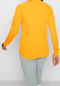 Top de manga larga naranja brillante con dobladillo redondeado, que presenta una textura suave y un diseño ajustado. Combinado con pantalones gris claro.