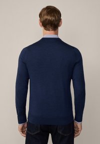 Maglione lavorato a maglia blu navy con maniche lunghe, polsini a coste e una finitura testurizzata. Indossato sopra una camicia con colletto, che mostra un dettaglio a strisce blu ai polsini.