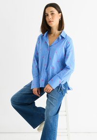 Frau, die auf einem weißen Hocker sitzt, trägt ein blau gestreiftes Hemd mit pinken Schleifen und weite blaue Jeans vor einem schlichten weißen Hintergrund.