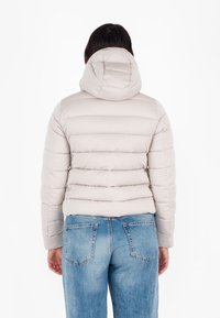 Lichtbeige pufferjack met capuchon, gewatteerd ontwerp en cropped pasvorm. Draagt over blauwe denim jeans, wat een casual stijl toont.