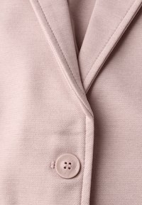 Tissu de blazer rose clair avec un tissage texturé, doté d'une fermeture à un seul bouton. Les détails remarquables incluent un col cranté et une coupe ajustée.