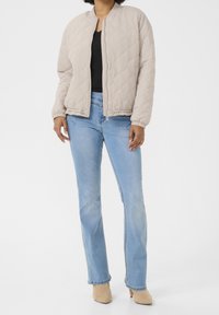 Person klädd i en beige vadderad jacka, svart topp, ljusblå jeans och beige klackstövlar som står emot en vit bakgrund.