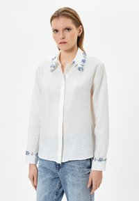 Camicia bianca con tessuto testurizzato, con accenti floreali blu ricamati sul colletto e sui polsini, maniche lunghe e chiusura con bottoni davanti.
