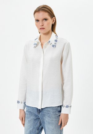 Witte blouse van gestructureerde stof, geborduurde blauwe bloemenaccenten op de kraag en manchetten, lange mouwen en een knoopsluiting aan de voorkant.