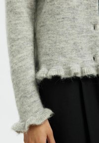 Jasnoszary, puszysty rękaw swetra i dół z falbankami, oparty o czarne spodnie, ukazujący część dłoni.