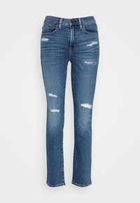 Blå denimjeans med en smal passform, som har flera slitna lappar och en medelhög midja. Klassisk femfickdesign och knäppning.