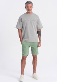 T-shirt gris à manches courtes avec le texte "Soyez le changement", associé à un short vert. Le modèle porte des baskets blanches, mettant en avant une tenue décontractée.