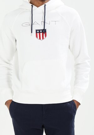 Biała bluza z kapturem z logo GANT i emblematem tarczy z gwiazdami i paskami, noszona z ciemnoniebieskimi spodniami, ręce w kieszeniach, widoczny tułów.