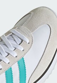 Detailný záber bielej tenisky s béžovými semišovými vrstvami na špičke, bielymi šnúrkami a tyrkysovými cik-cak pruhmi na bočnej strane.