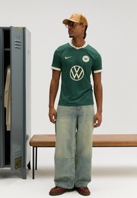 Zöld Nike mez bézs szegéllyel és Volkswagen logóval, széles világoskék farmernadrággal és barna, masszív cipőkkel párosítva, egy minimál stílusú öltözőben.